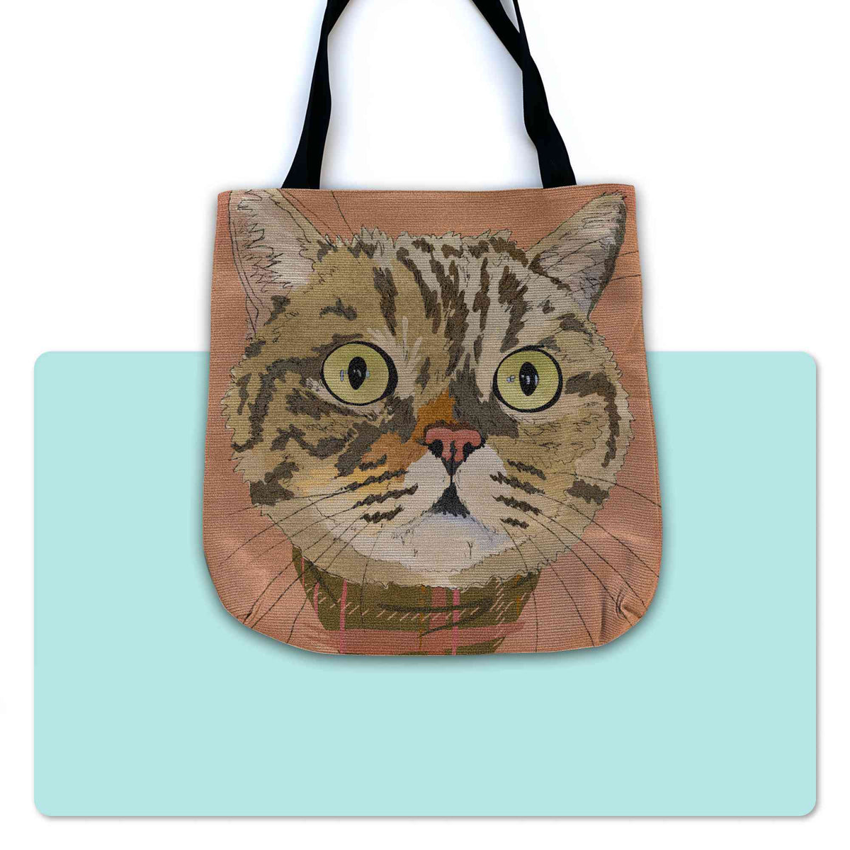 A woven tote bag with black shoulder straps featuring a custom illustrated cat portrait of a tabby cat wearing a plaid bandana set against a subtle textured orange background printed on the side of the bag, shown centered over a large light aqua rectangular shape on a white background.