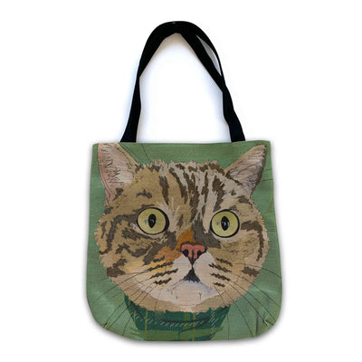 A woven tote bag with black shoulder straps featuring a custom illustrated cat portrait of a tabby cat wearing a plaid bandana set against a subtle textured green background printed on the side of the bag, shown on a clean white background.