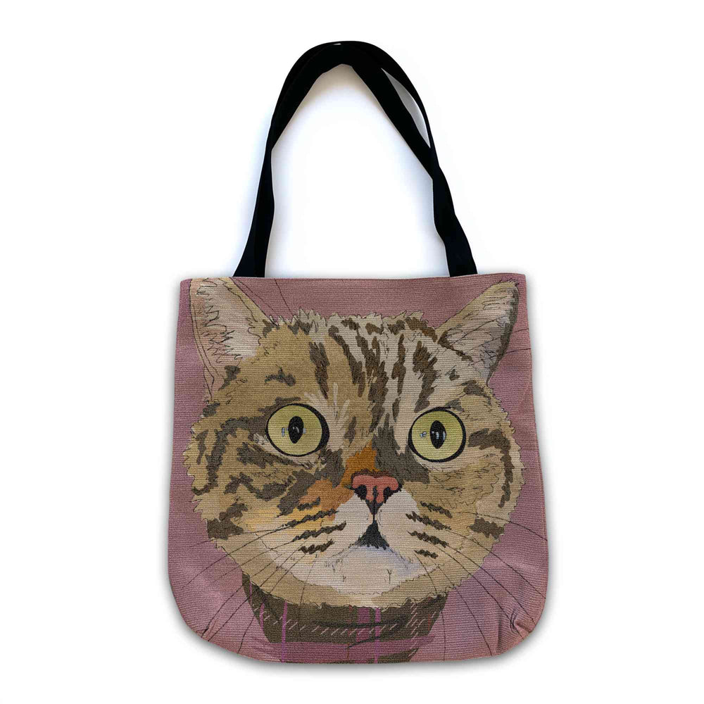 A woven tote bag with black shoulder straps featuring a custom illustrated cat portrait of a tabby cat wearing a plaid bandana set against a subtle textured dusty rose background printed on the side of the bag, shown on a clean white background. 