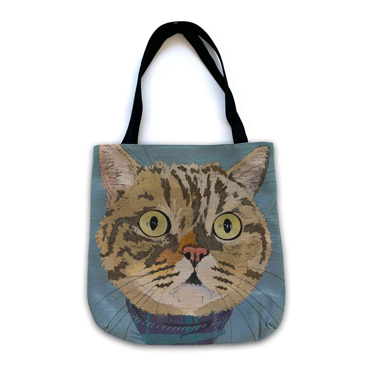 A woven tote bag with black shoulder straps featuring a custom illustrated cat portrait of a tabby cat wearing a plaid bandana set against a subtle textured blue background printed on the side of the bag, shown on a clean white background. 