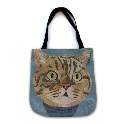A woven tote bag with black shoulder straps featuring a custom illustrated cat portrait of a tabby cat wearing a plaid bandana set against a subtle textured blue background printed on the side of the bag, shown on a clean white background. 