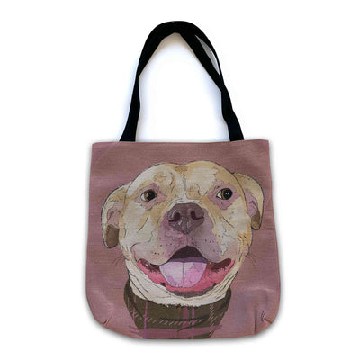 A woven tote bag with black shoulder straps featuring a custom illustrated dog portrait of a smiling tan dog wearing a plaid bandana set against a subtle textured dusty rose background printed on the side of the bag, shown on a clean white background.