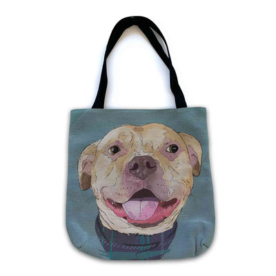 A woven tote bag with black shoulder straps featuring a custom illustrated dog portrait of a smiling tan dog wearing a plaid bandana set against a subtle textured blue background printed on the side of the bag, shown on a clean white background. 