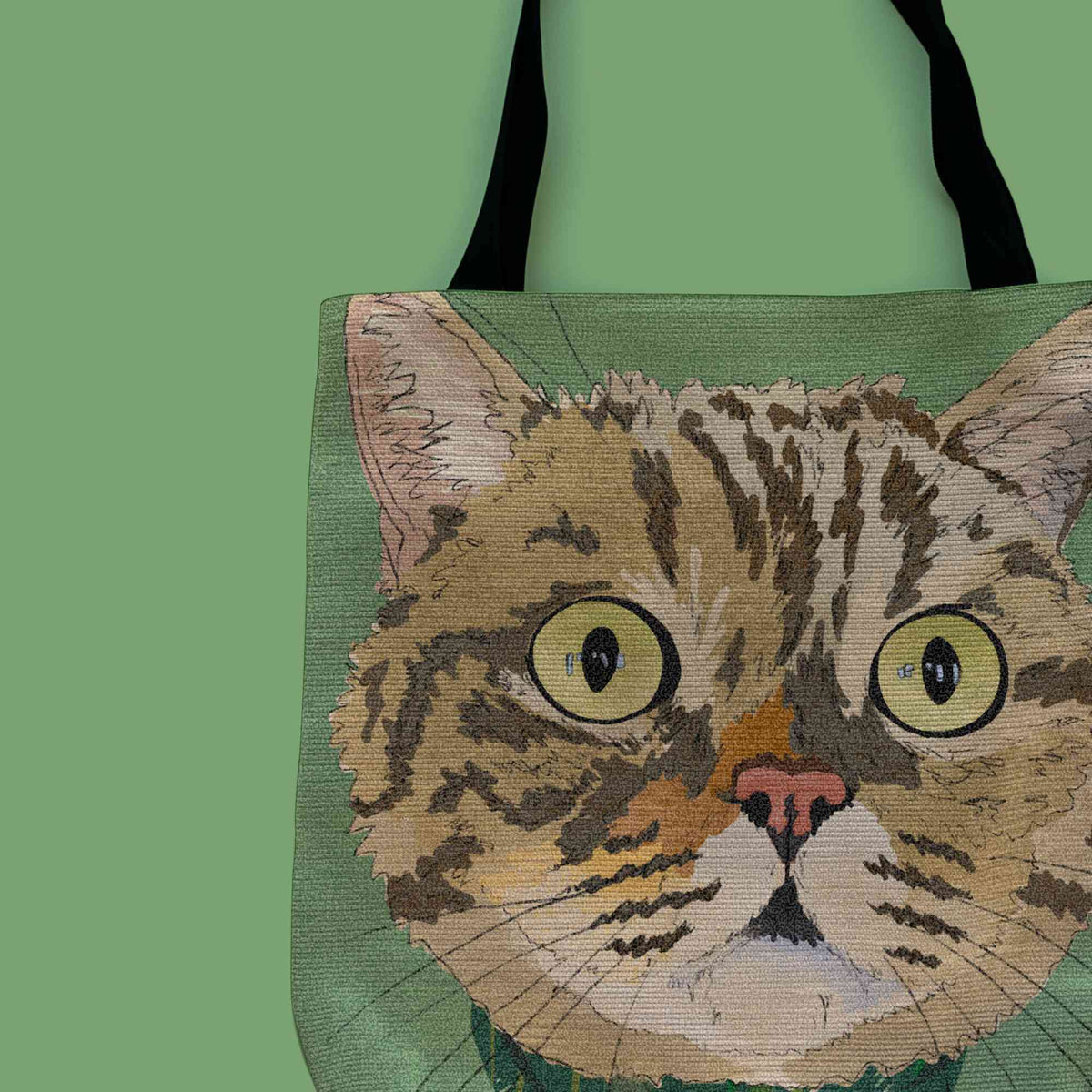 A close-up woven tote bag with black shoulder straps featuring a custom illustrated cat portrait of a tabby cat wearing a plaid bandana set against a subtle textured green background printed on the side of the bag, shown on a green background. 